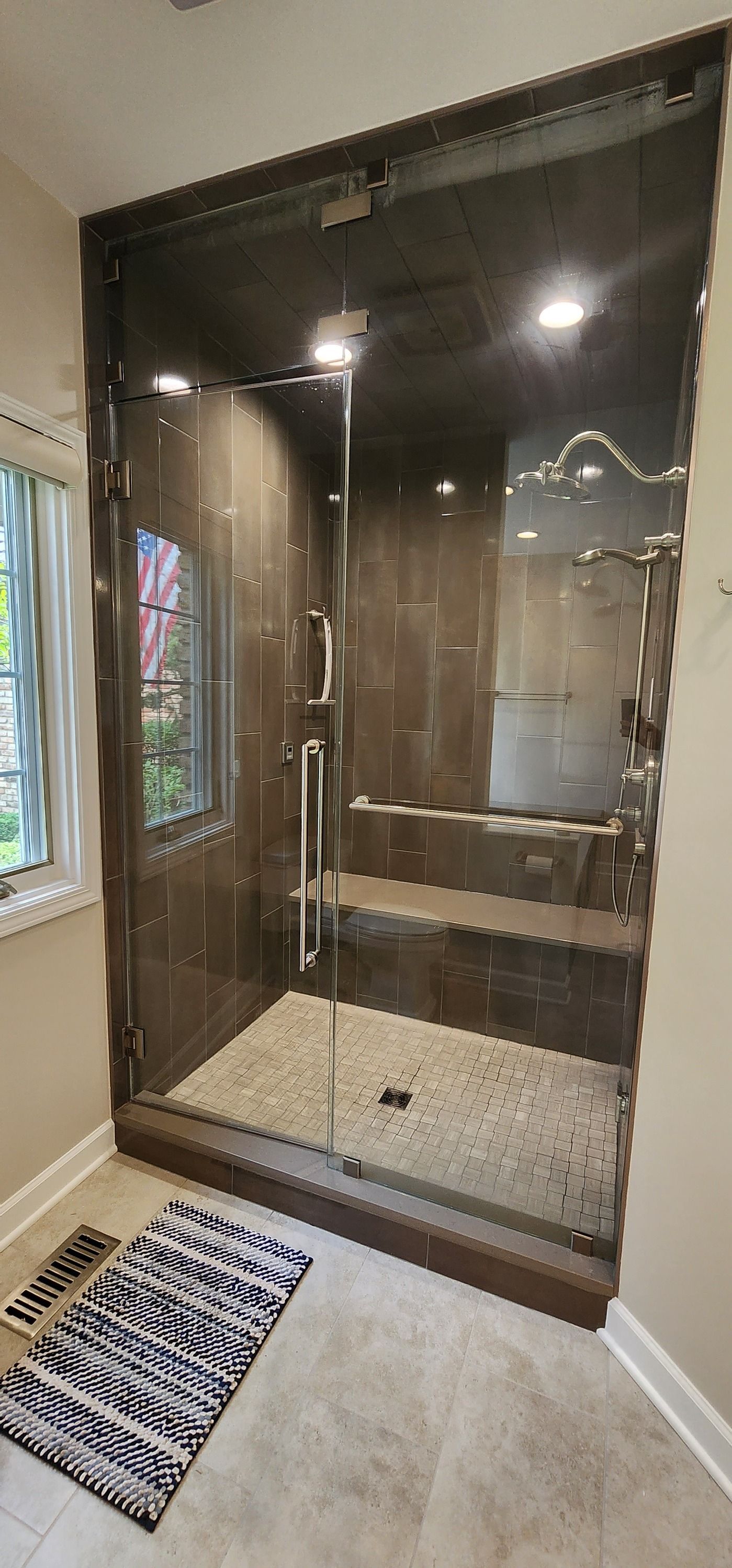 A walk-in shower with a glass door, brown tile, and a bench. A black and white rug is in front.