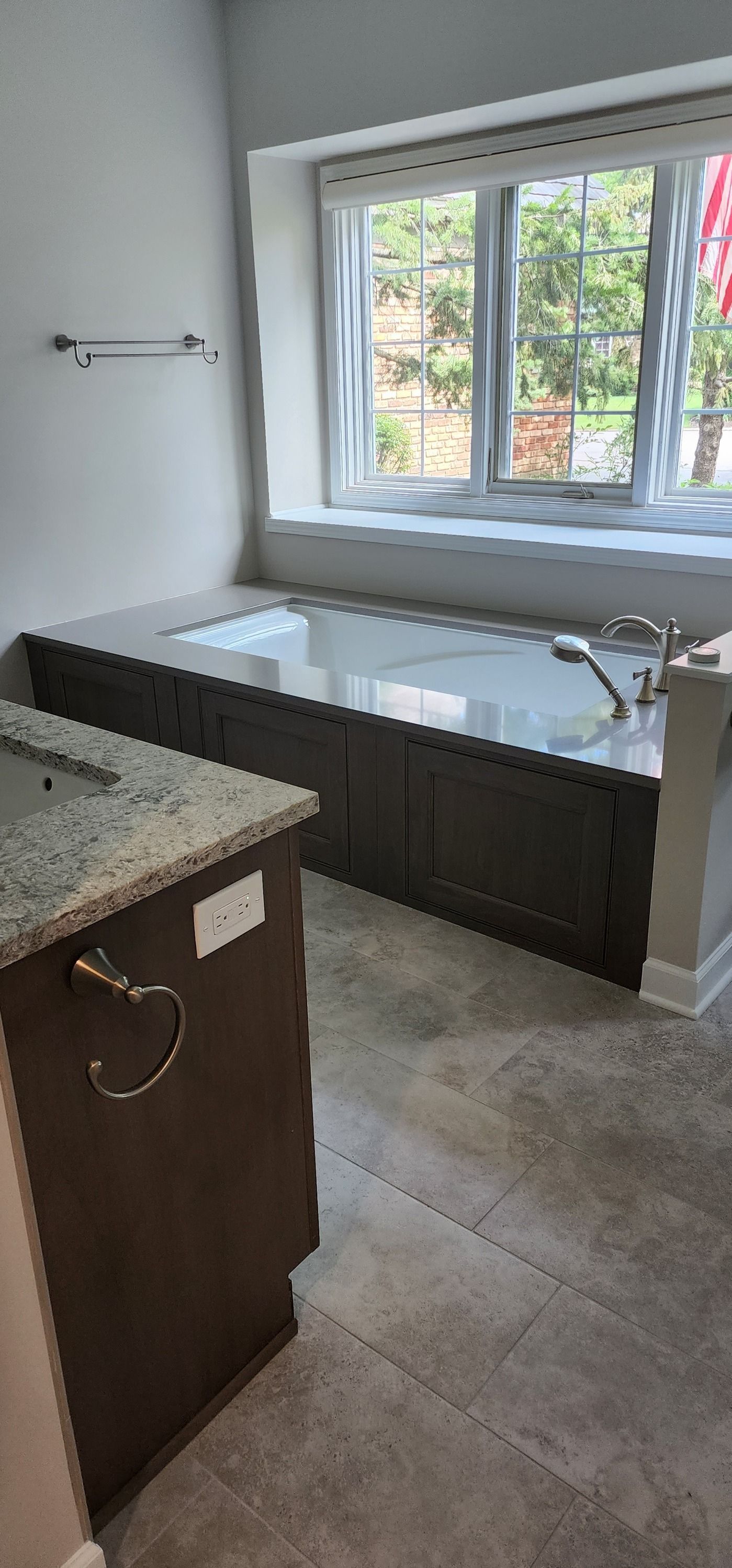 A bathroom with a built-in tub under a window. Dark cabinets and gray tile.