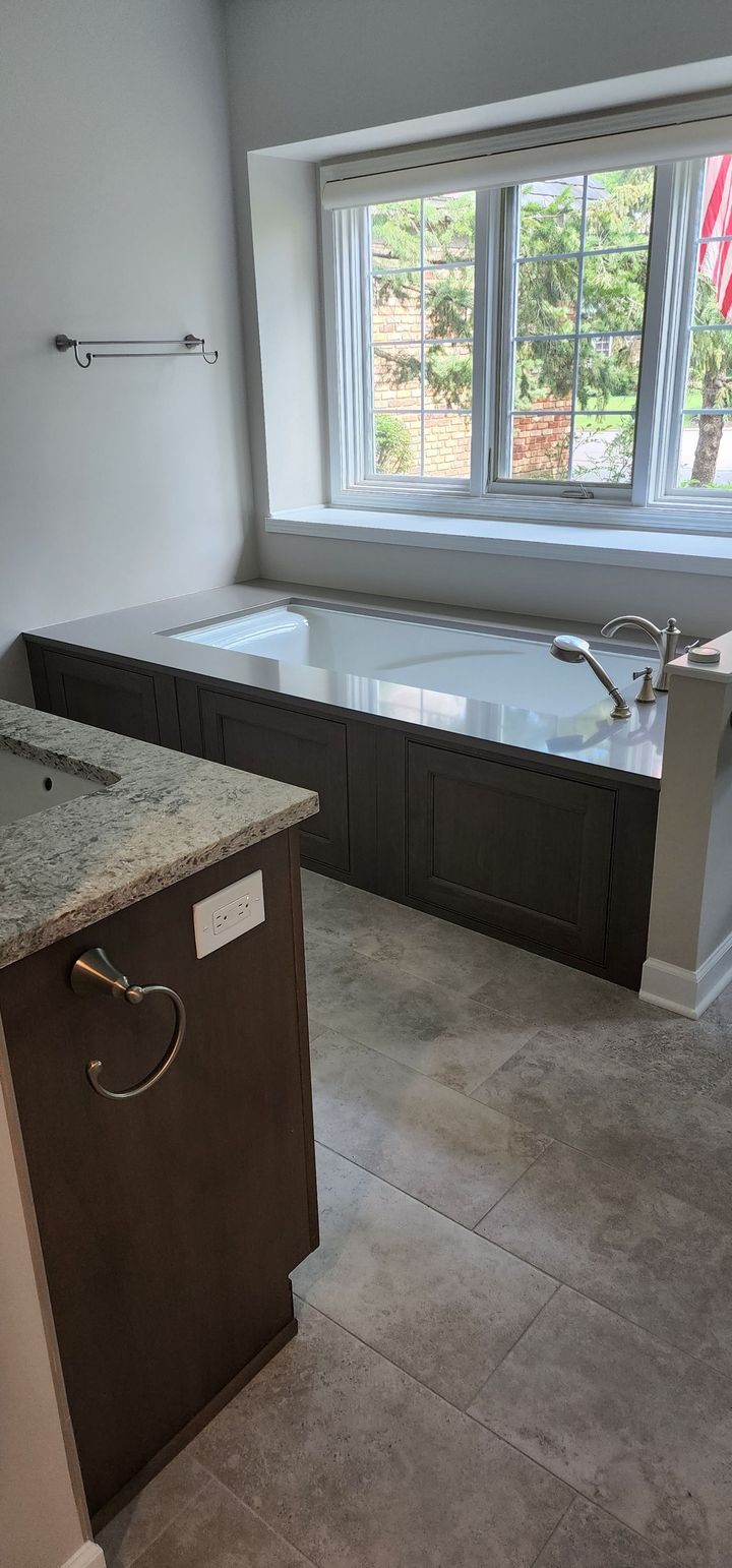 A bathroom with a built-in tub under a window. Dark cabinets and gray tile.