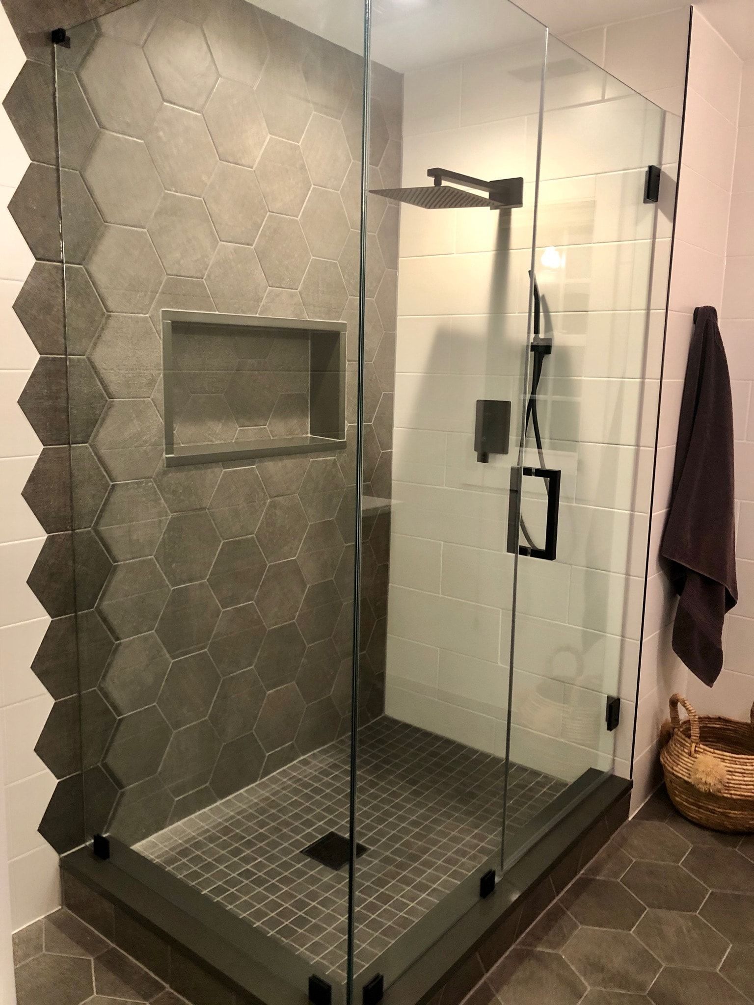 Modern shower with gray hexagon tiles, glass door, and dark fixtures. A towel hangs on the right.