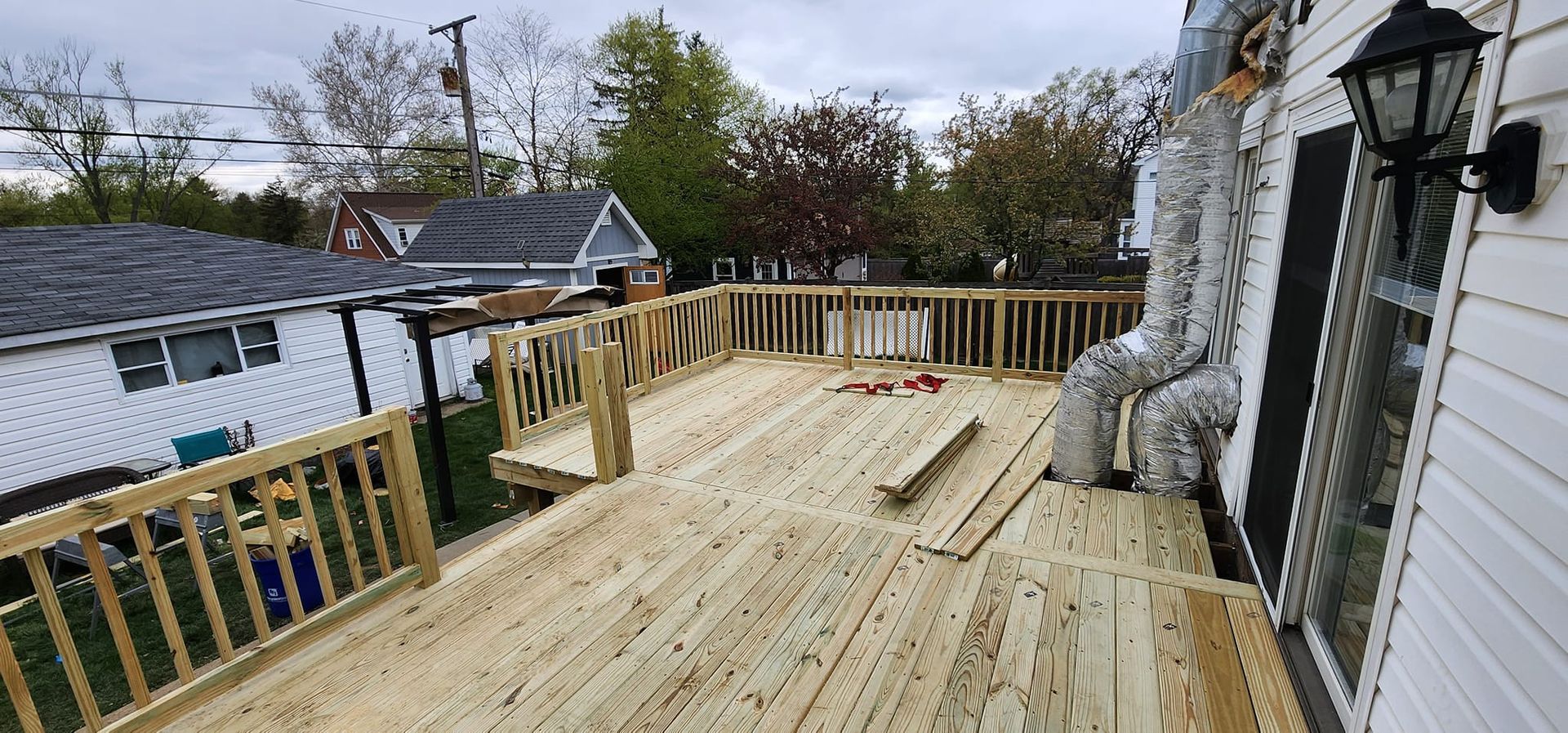 Wooden deck with railings, next to a white house with trees in the background.