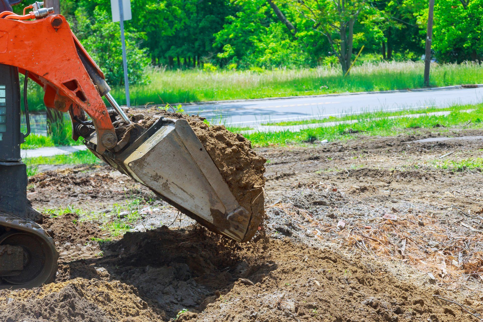 Orange excavator bucket dumping dirt onto a grassy area near a road.