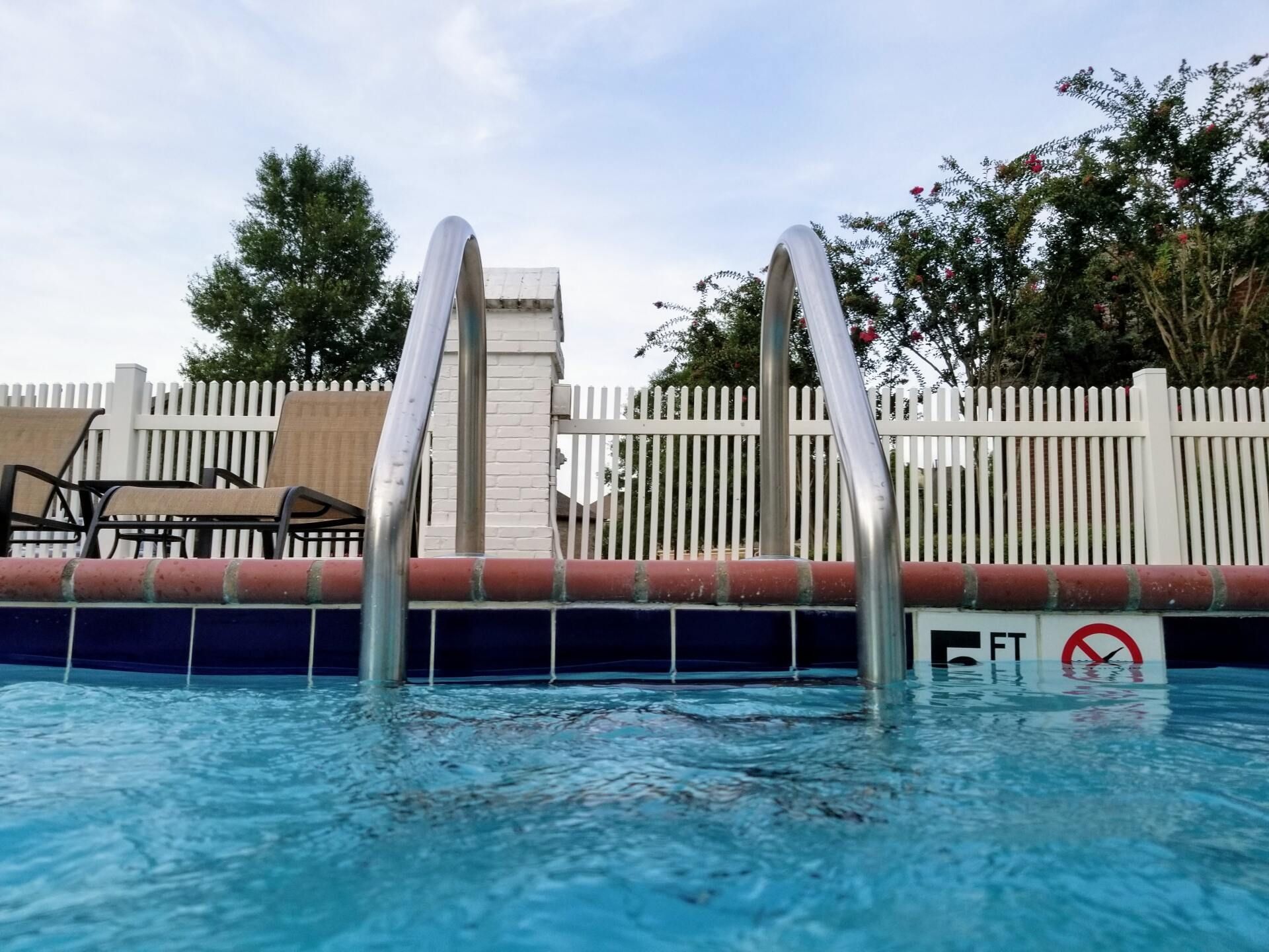Pool ladder with stainless steel handrails against a blue pool. White fence and trees in background.