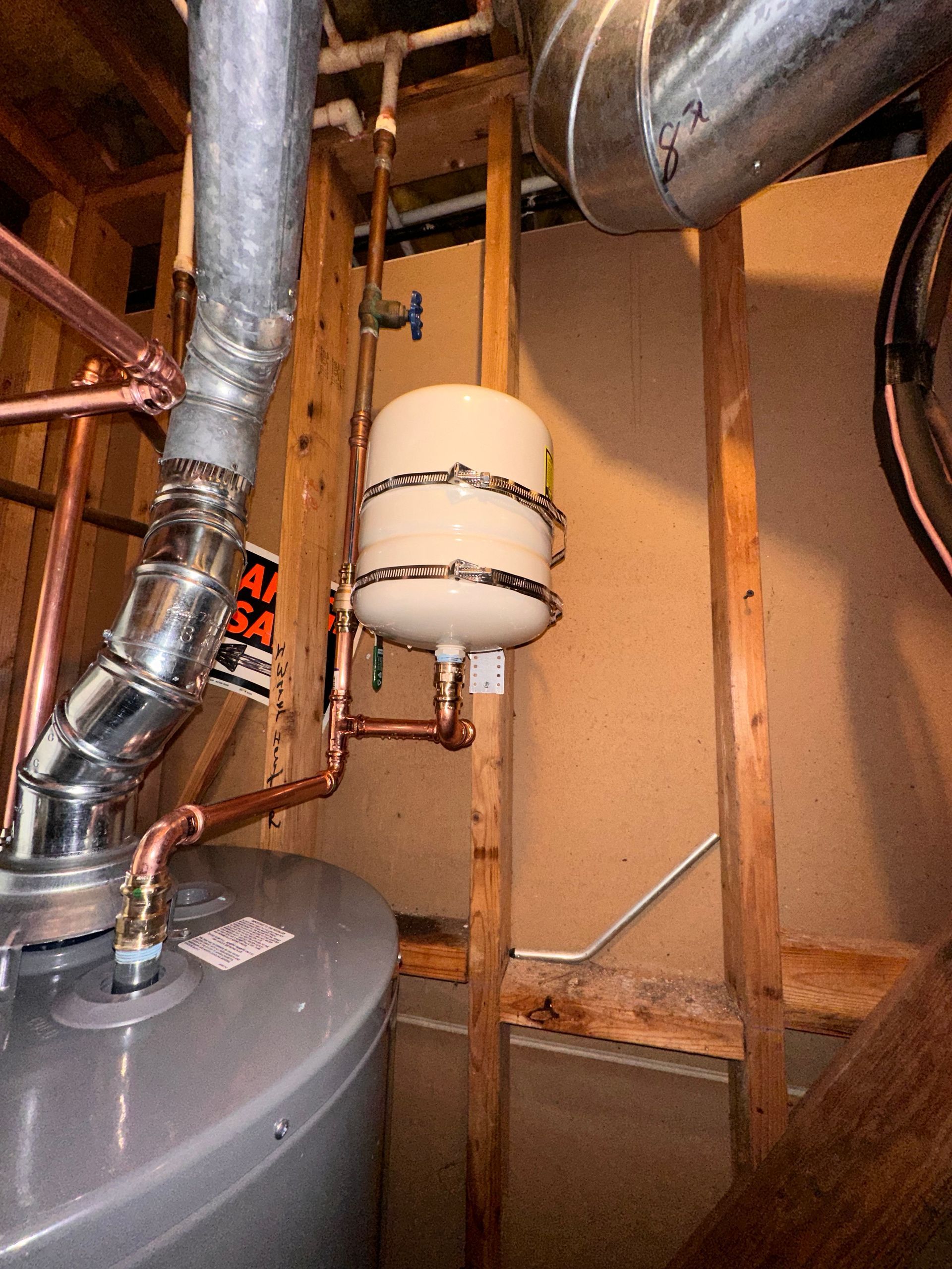Water heater with a small, cylindrical expansion tank, copper pipes, and metal ductwork in a utility room.