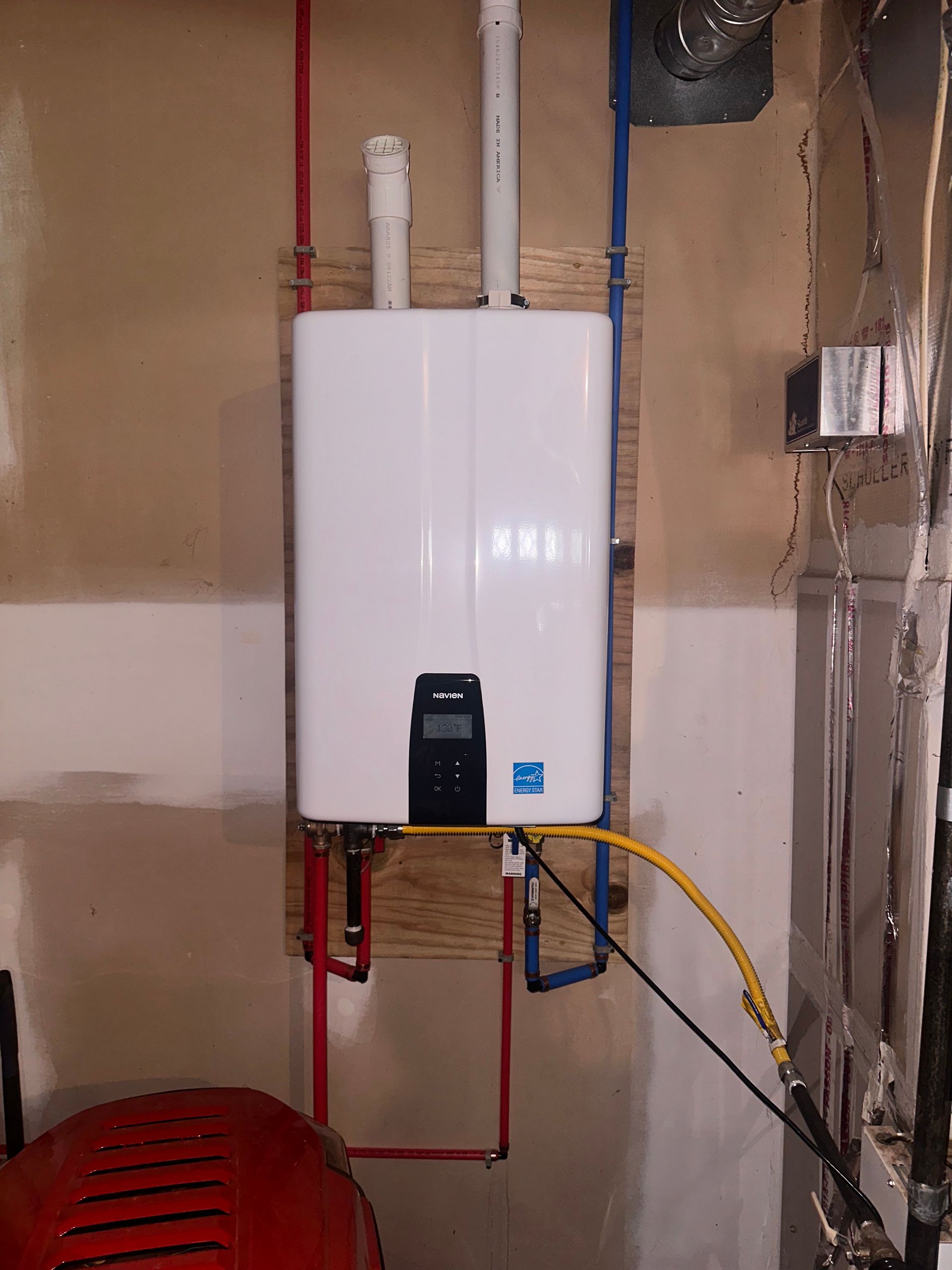 White tankless water heater mounted on a wooden board against a beige wall, with connected pipes and wiring.
