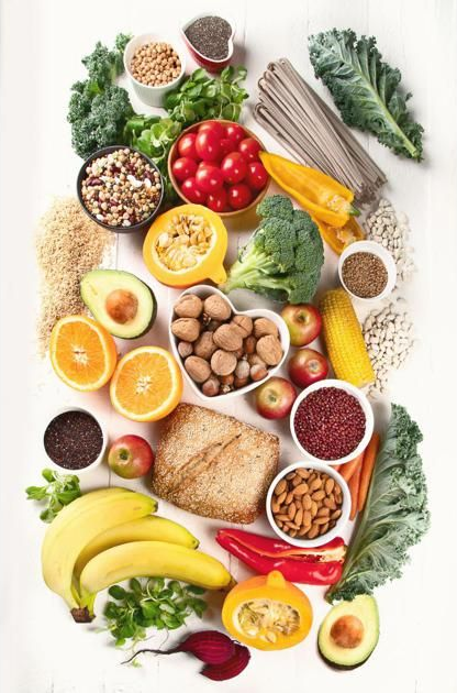 A flat lay of various healthy foods including vegetables, fruits, nuts, grains, and whole-wheat bread on a white surface.