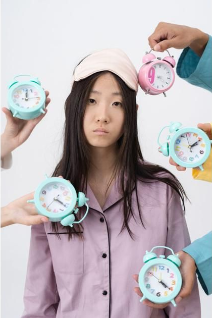 A tired-looking person in pajamas with a sleep mask is surrounded by hands holding multiple alarm clocks.