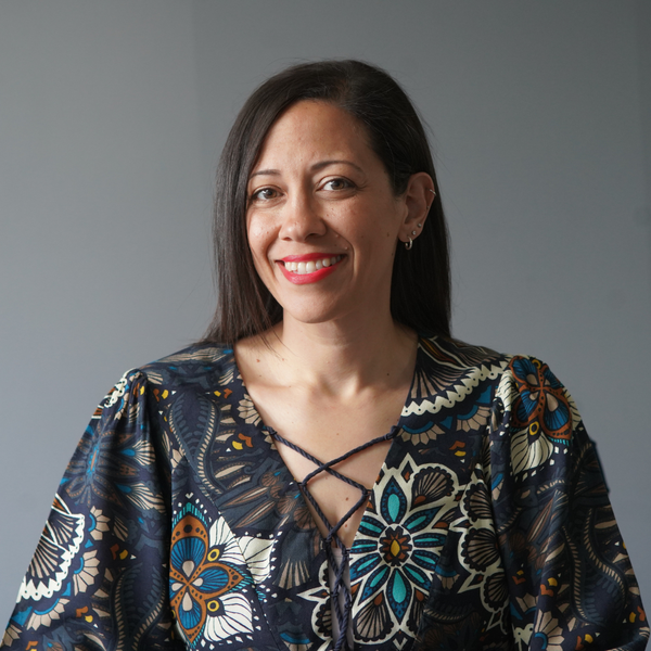 A person with dark hair smiling at the camera, wearing a dark blue, patterned floral top against a gray background.