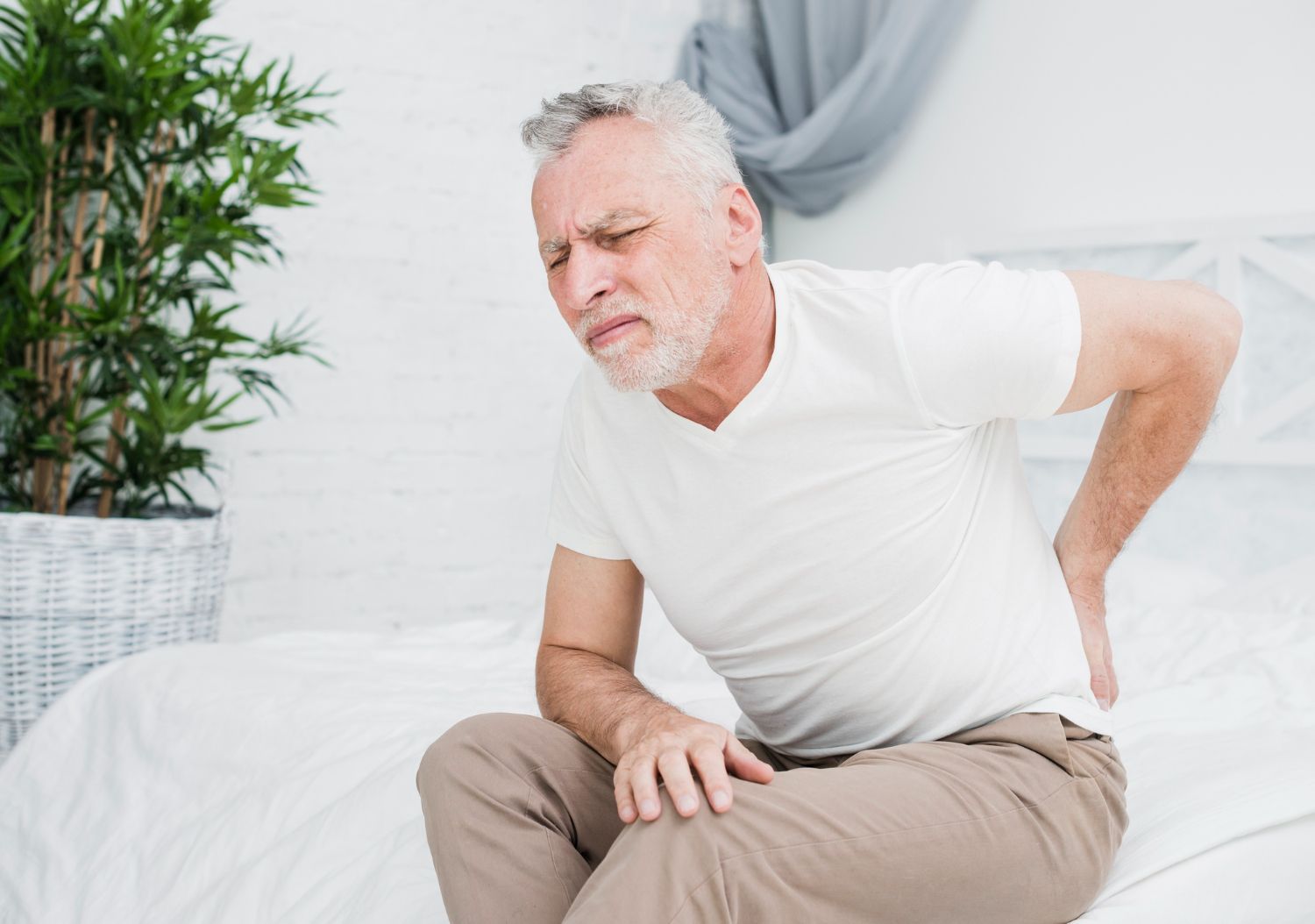 A person sitting on a bed, grimacing in pain while holding their lower back.
