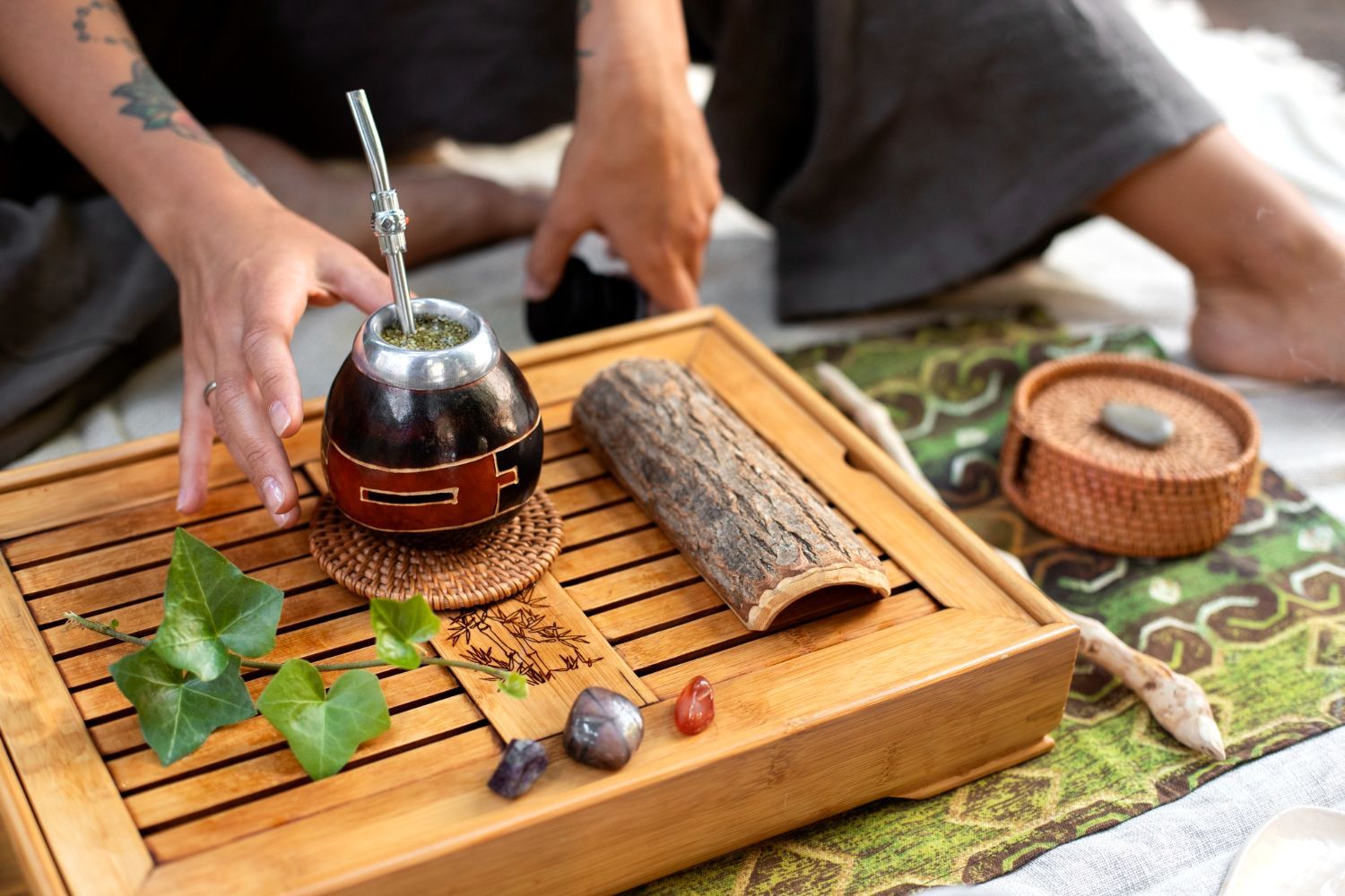 A person prepares yerba mate on a wooden tray with ivy leaves, a piece of bark, and smooth stones.