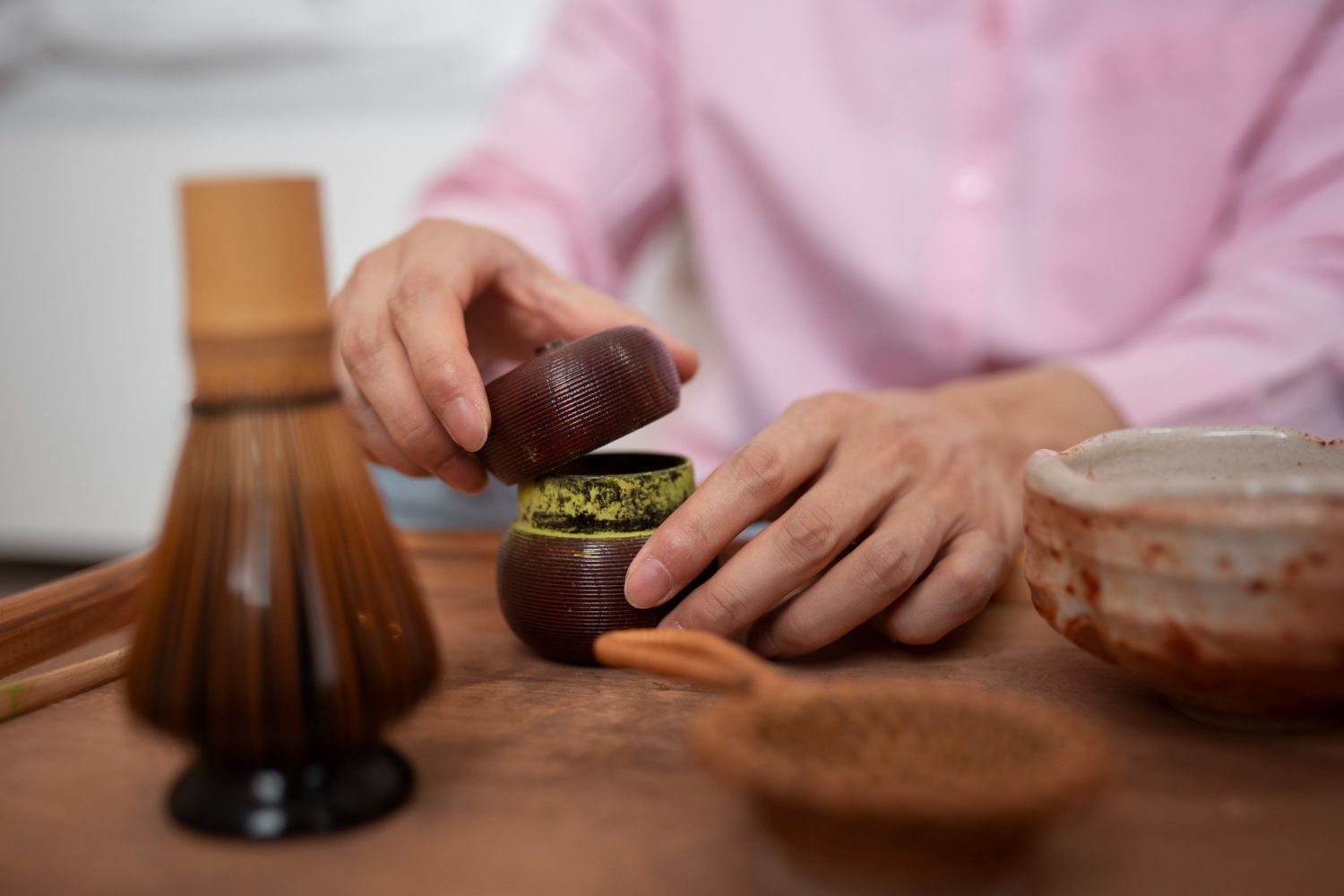 Hands open a small, ribbed dark wooden tea caddy filled with green matcha powder on a wooden tray with a bamboo whisk.