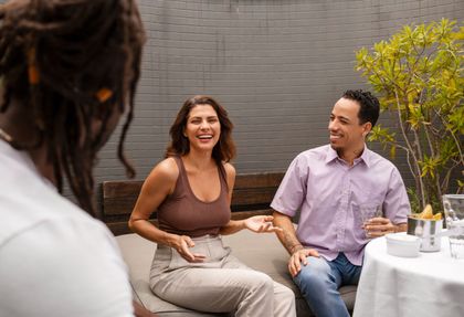 Three people sitting on an outdoor patio, two facing each other while laughing and gesturing, with a third person blurred.