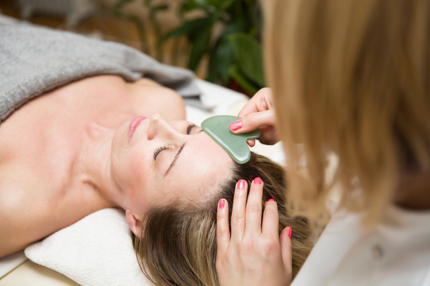 A practitioner uses a green gua sha stone tool to perform a facial massage on a person lying down in a spa setting.