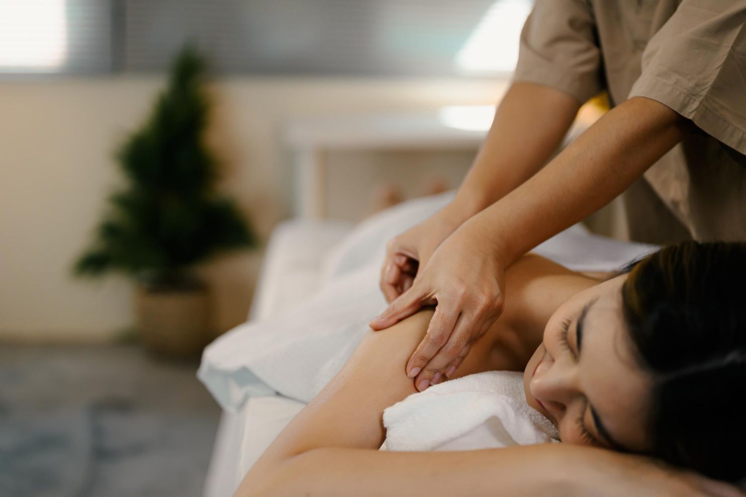 A person receives a shoulder massage from a professional in a peaceful, softly lit room with a plant in the background.