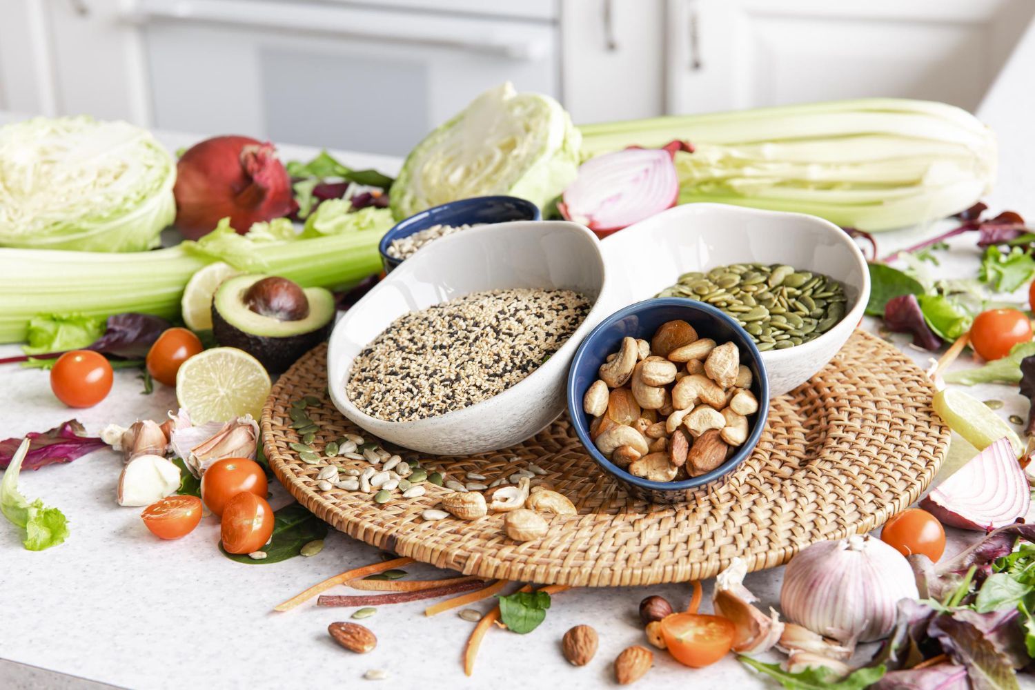 A selection of fresh vegetables, nuts, seeds, and grains arranged on a table around bowls of cashews, seeds, and quinoa.