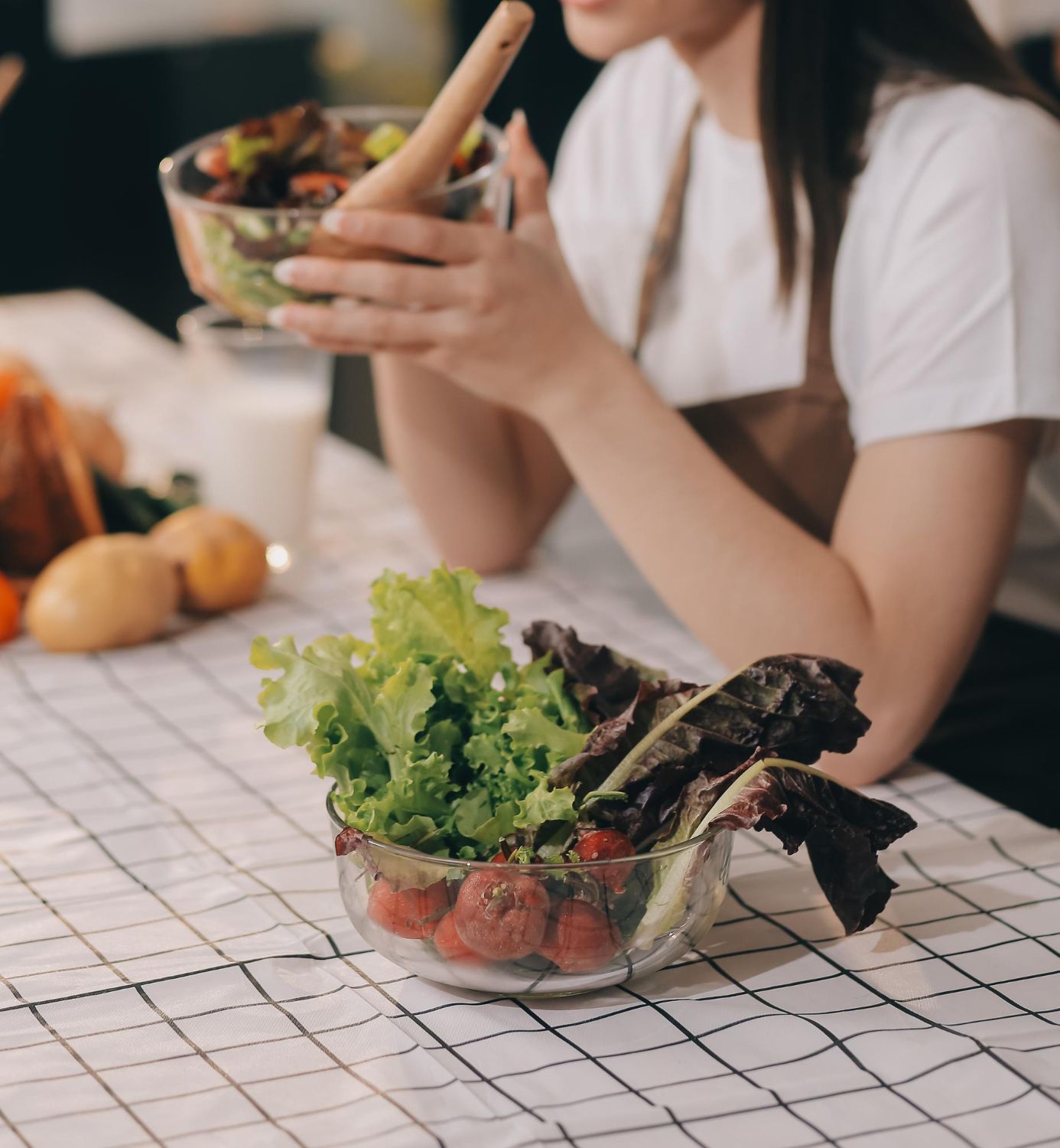 A bowl of fresh salad sits on a grid-patterned tablecloth while a person prepares another bowl of salad in the background.