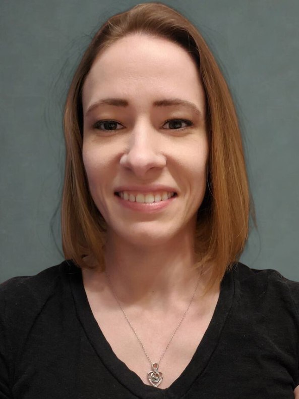 A person with shoulder-length brown hair and a silver heart pendant necklace smiling against a grey background.