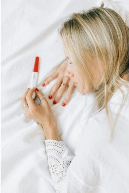 A person lies on a white bed, holding a pregnancy test while looking down at it with a contemplative expression.