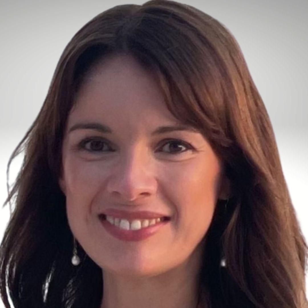 Woman with brown hair smiles, wearing a black top. Lit by sunlight with green plants in background.