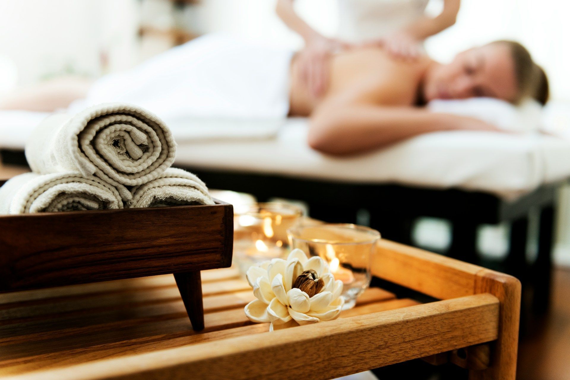 Rolled white towels and a flower on a wooden table, with a person receiving a massage in the blurred background.