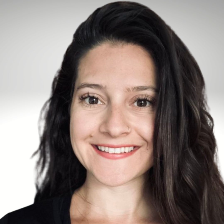 Woman with brown hair smiles, wearing a black top. Lit by sunlight with green plants in background.