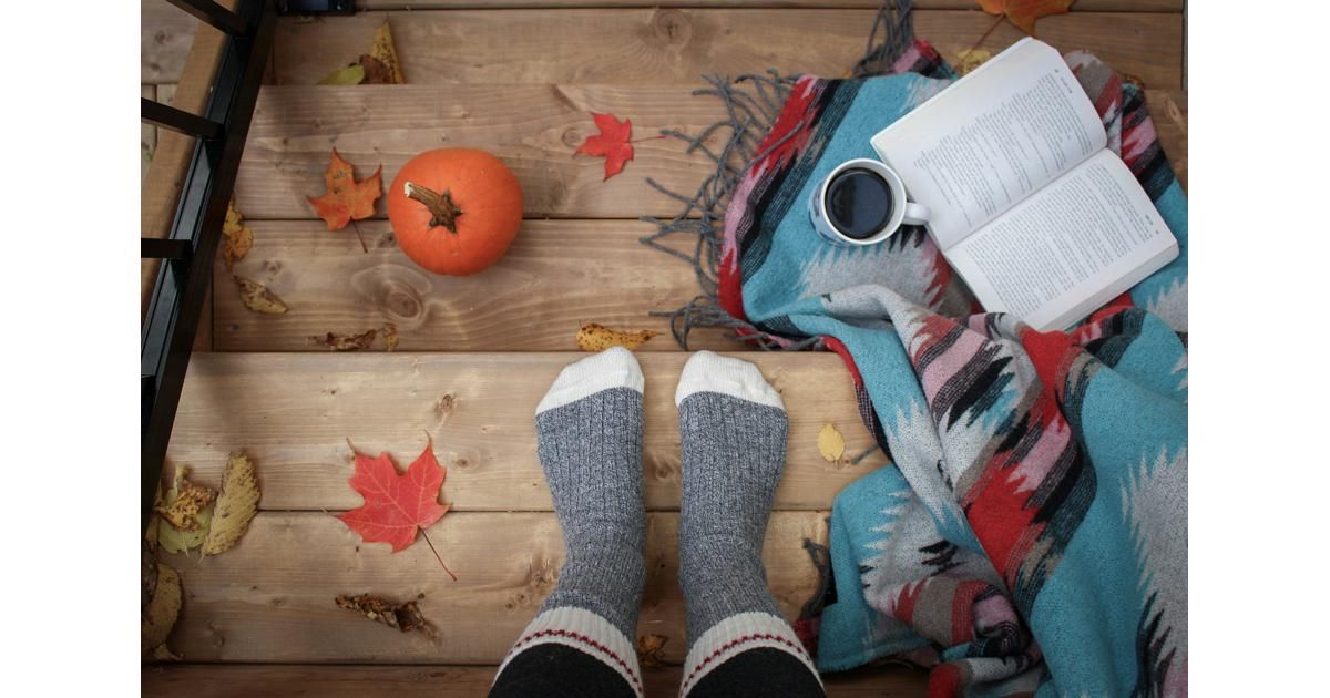 Feet in gray socks resting on wooden steps with a pumpkin, fallen leaves, a patterned blanket, and an open book.