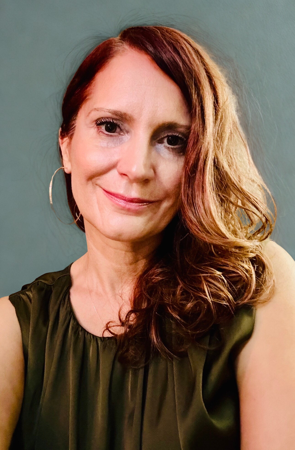 A person with long, wavy auburn hair and silver hoop earrings, wearing a sleeveless olive top against a gray background