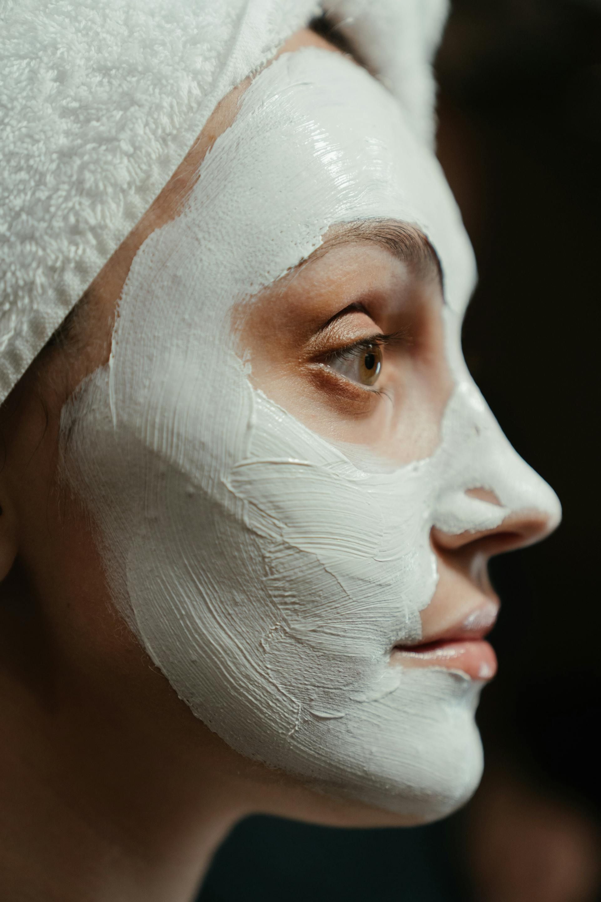 Profile view of a person wearing a white clay facial mask and a white towel wrapped around their head.