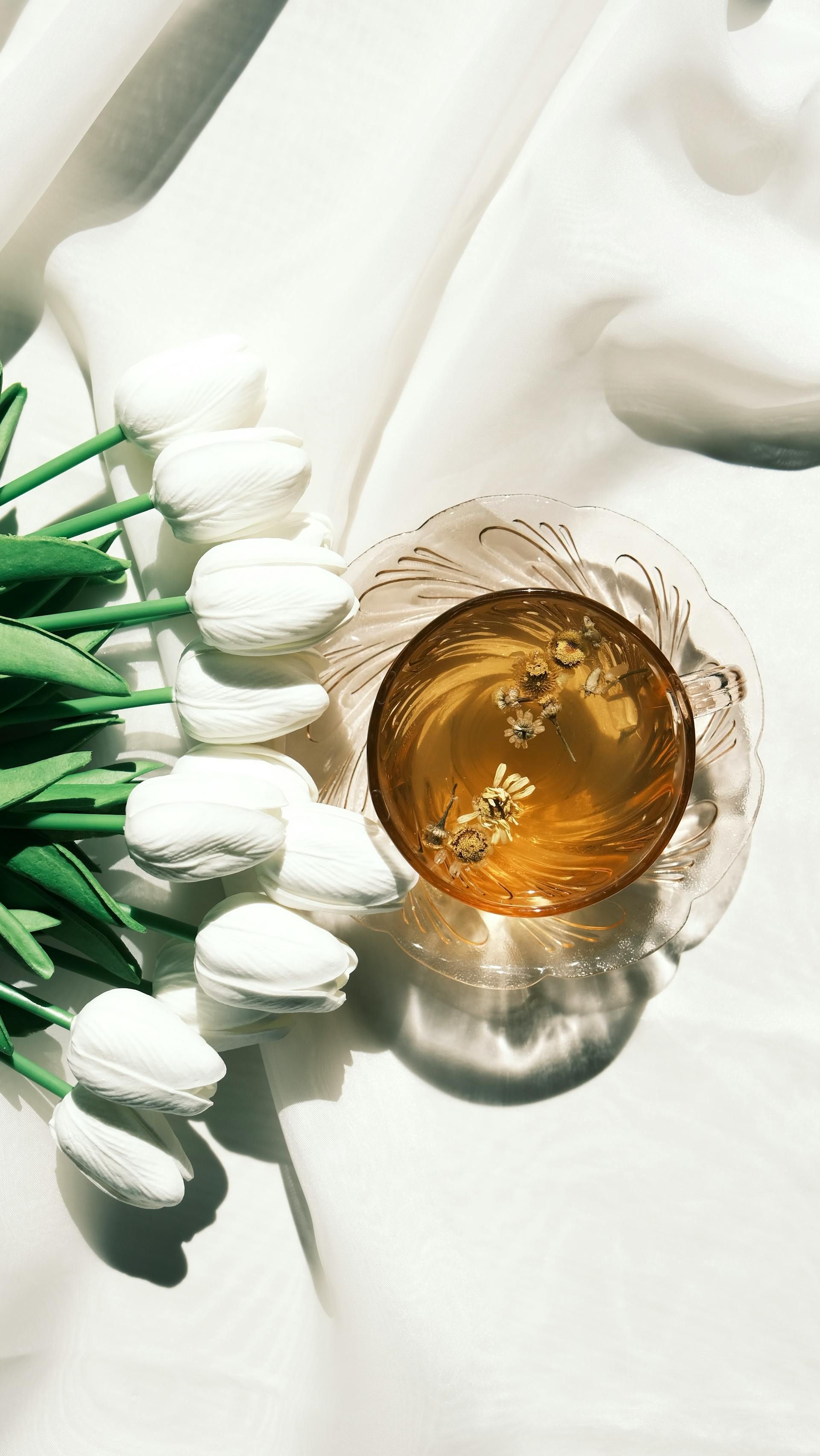 A cup of golden tea with floating flower buds sits on a white cloth next to a bunch of white tulips.