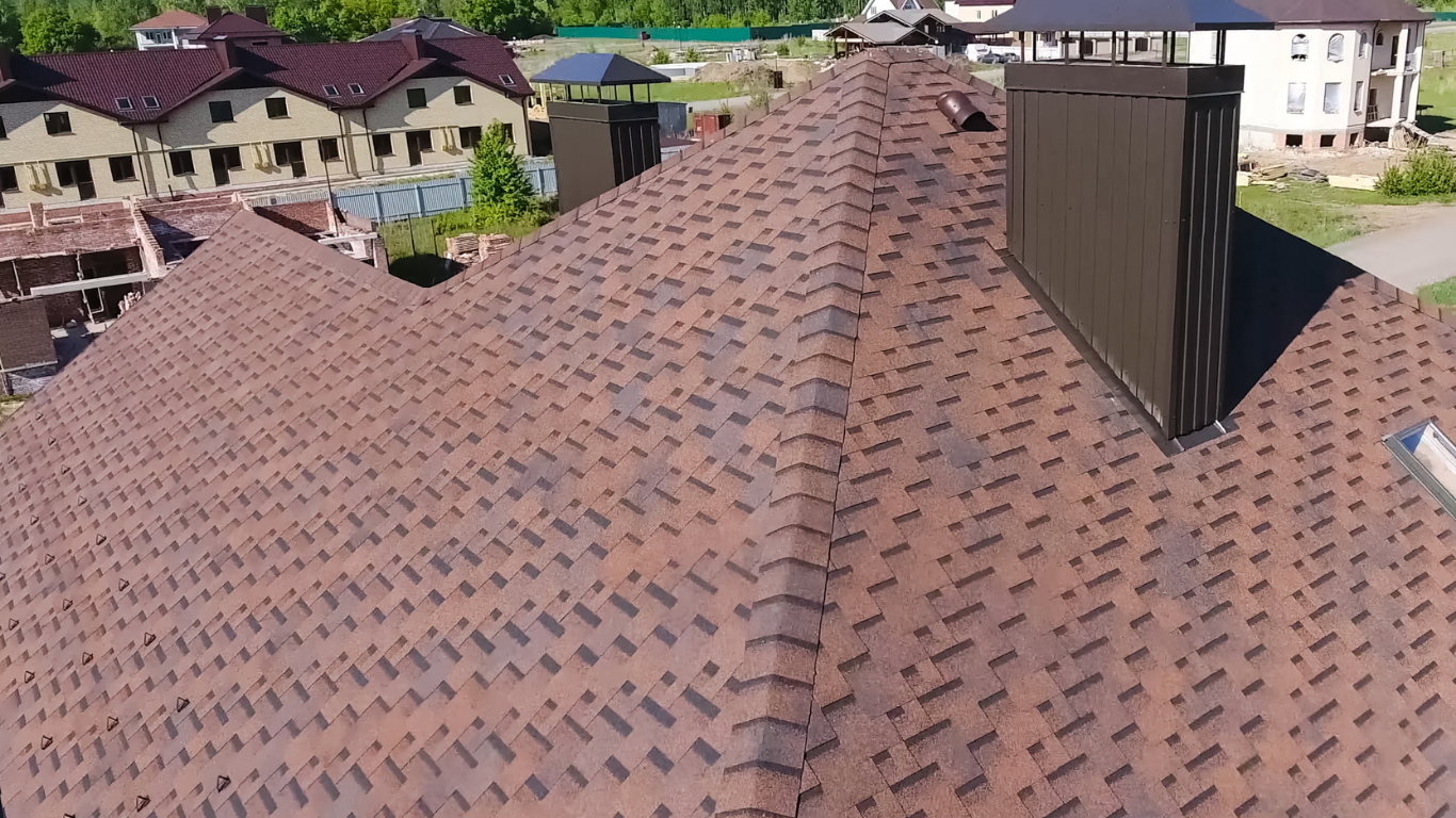 Aerial view of a brown shingled roof with a dark chimney in a suburban neighborhood