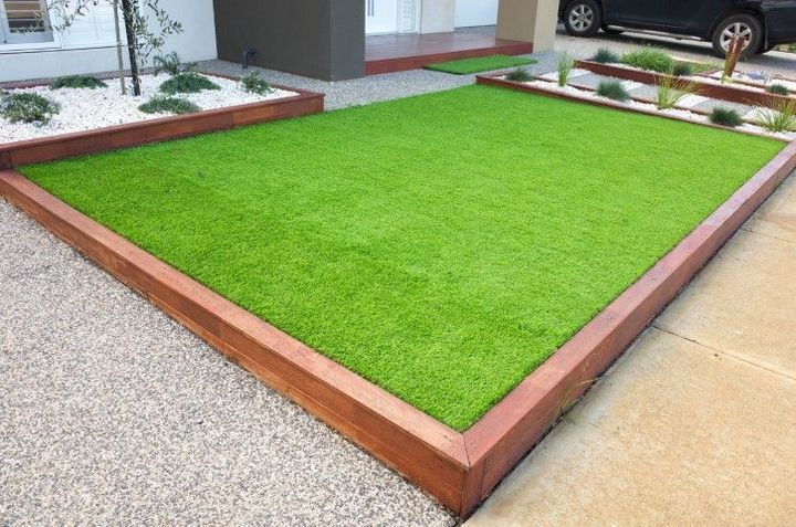 Raised square lawn with bright green artificial grass bordered by reddish wood next to a sidewalk and building
