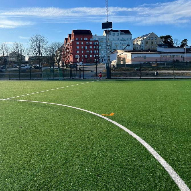 Green sports field with white lines, fenced in front of city buildings under a blue sky