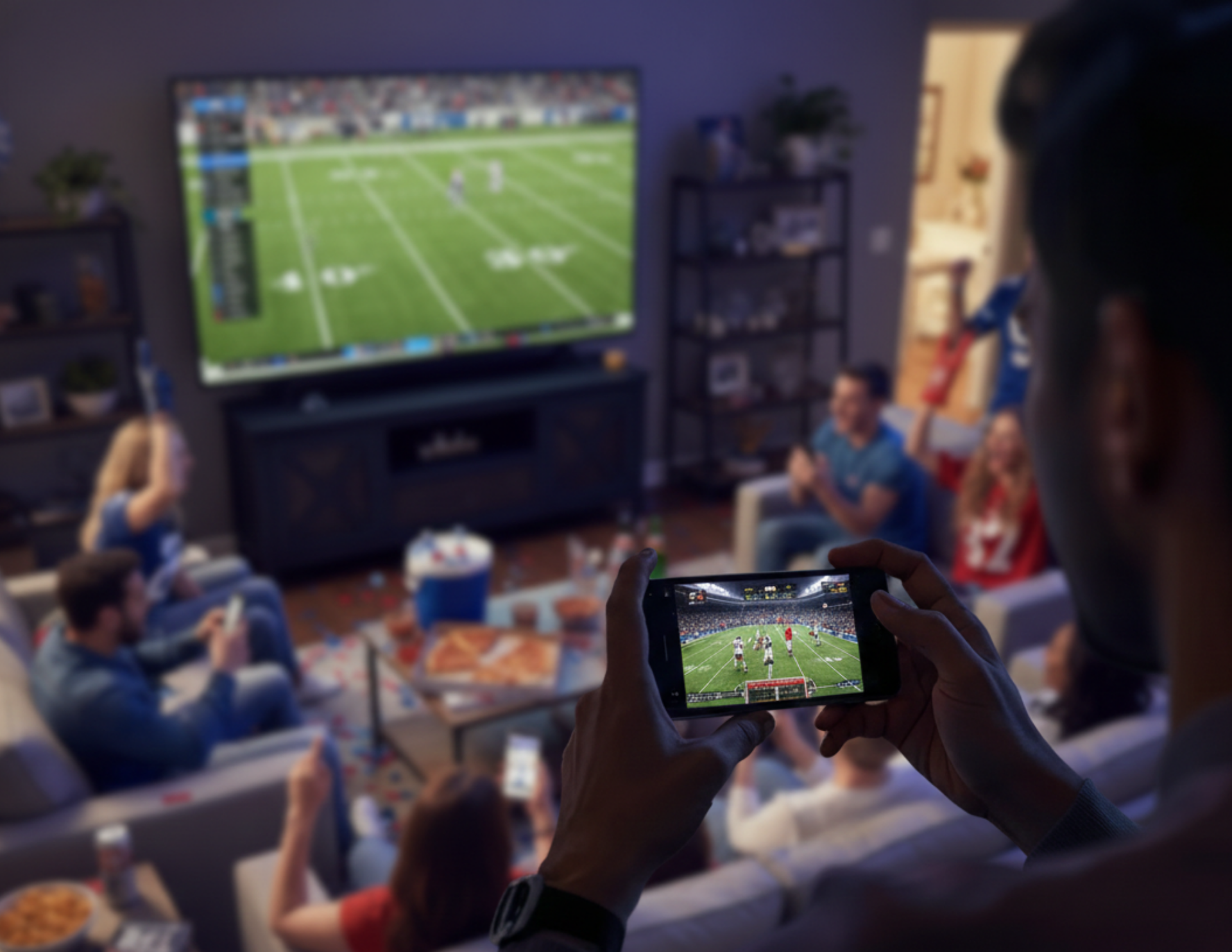 A person holds a phone showing a football game while friends watch the same game on a large TV