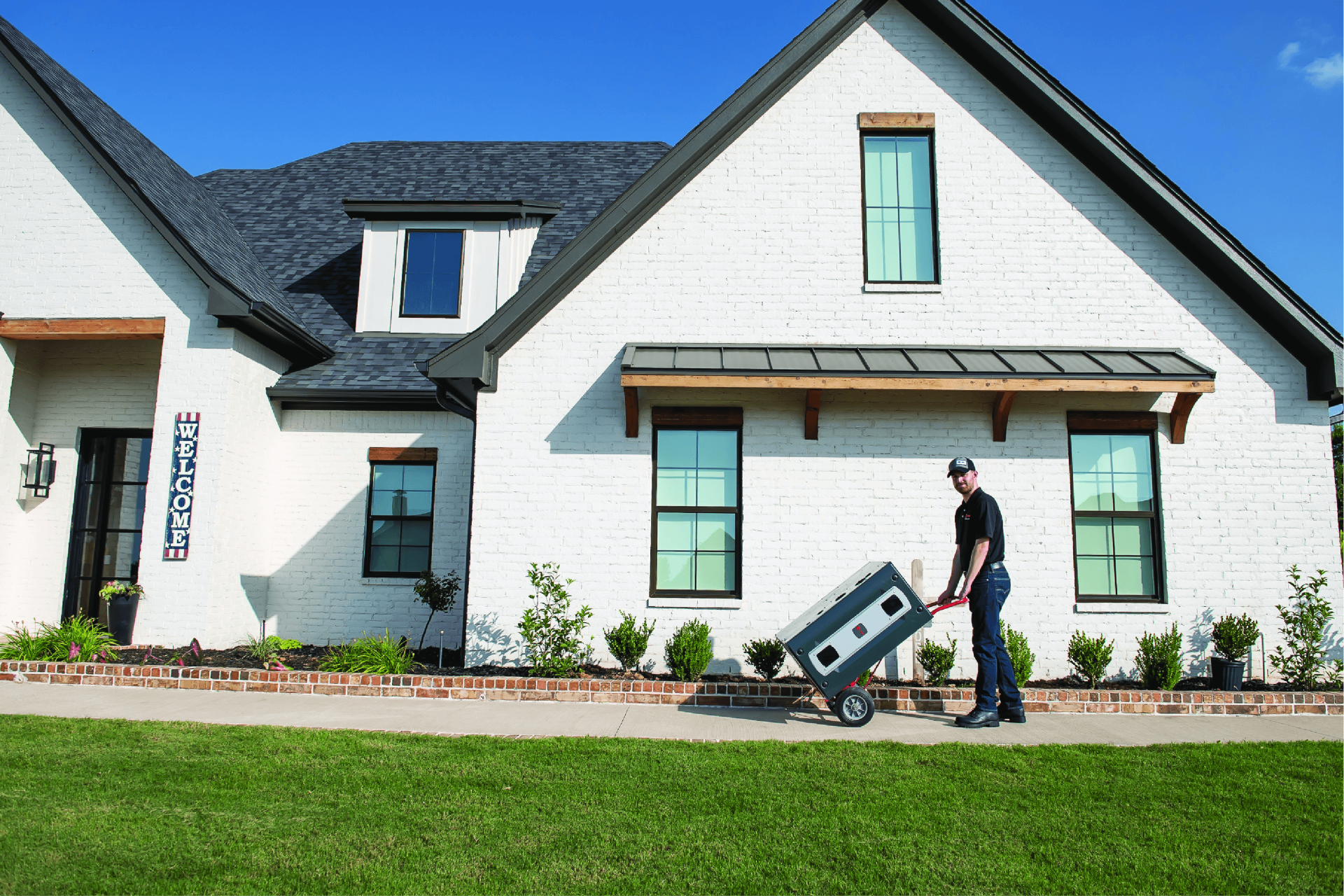 Man rolling a gray appliance on wheels on a sidewalk in front of a white brick house with a dark gray roof.