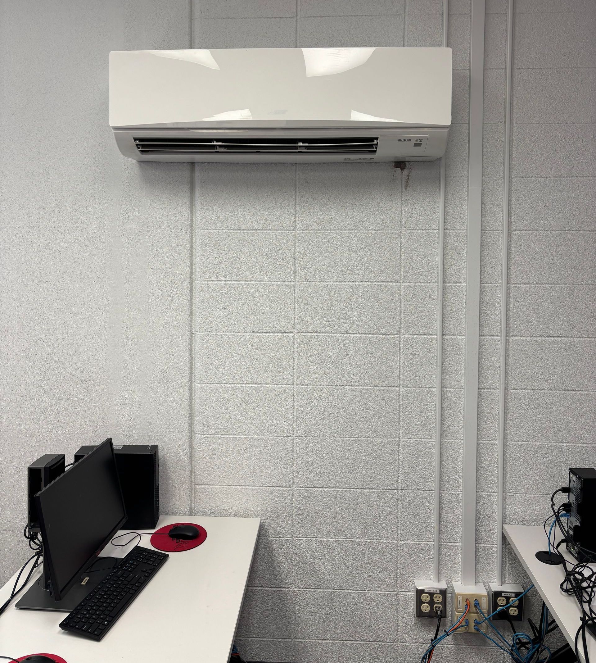 White air conditioner mounted on a textured gray wall above a desk with computer and speakers.