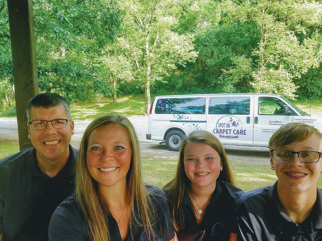 Family of four smiles outdoors; van in background, trees in background.