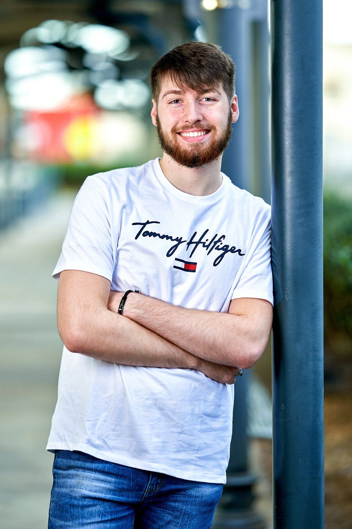 A young man with a beard is leaning against a pole with his arms crossed.
