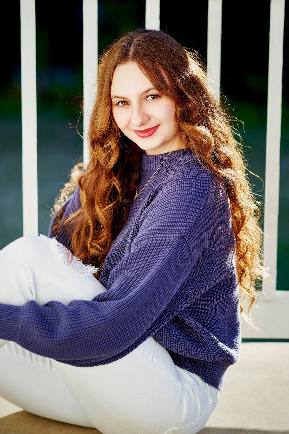 A young woman is sitting on the ground wearing a purple sweater and white pants.