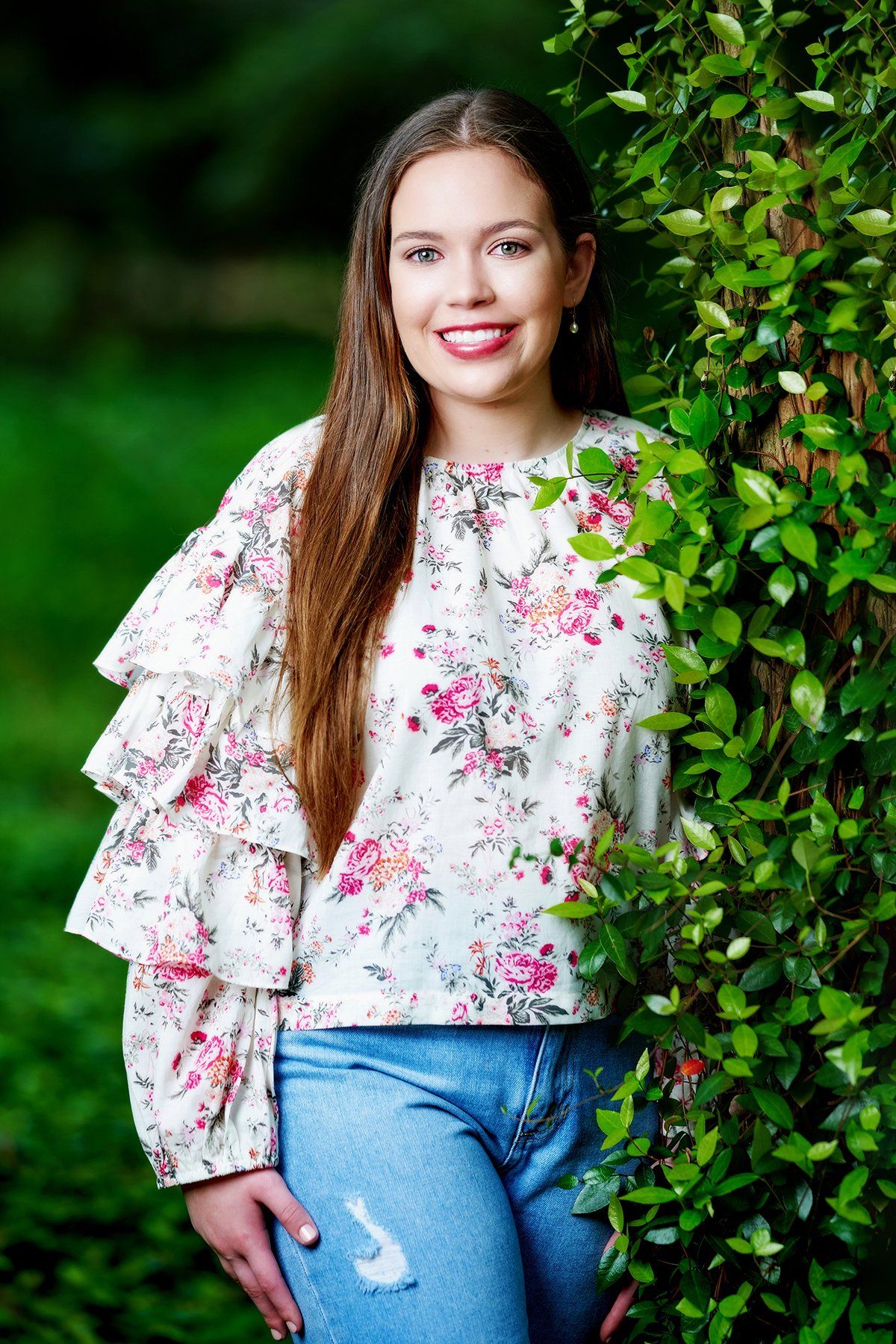 A young woman in a floral shirt and jeans is standing next to a tree.