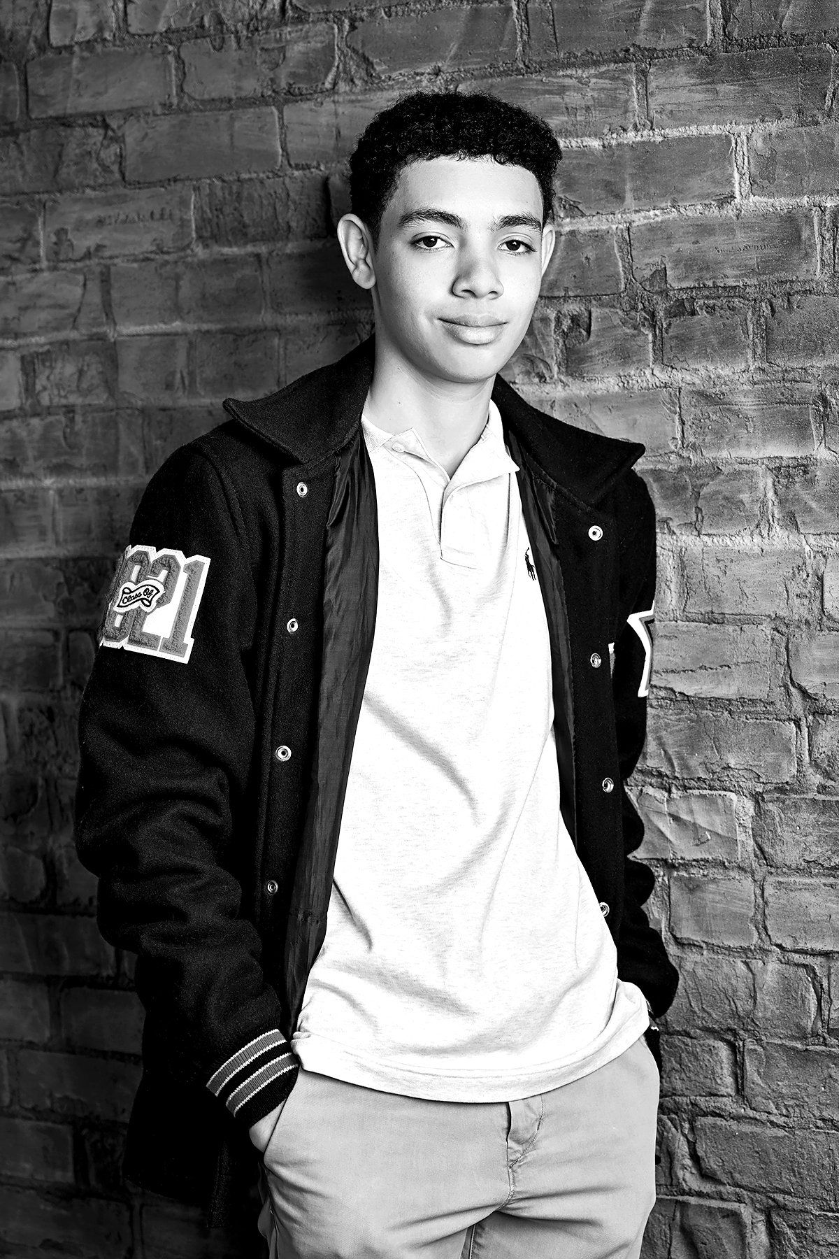 A black and white photo of a young man standing in front of a brick wall.