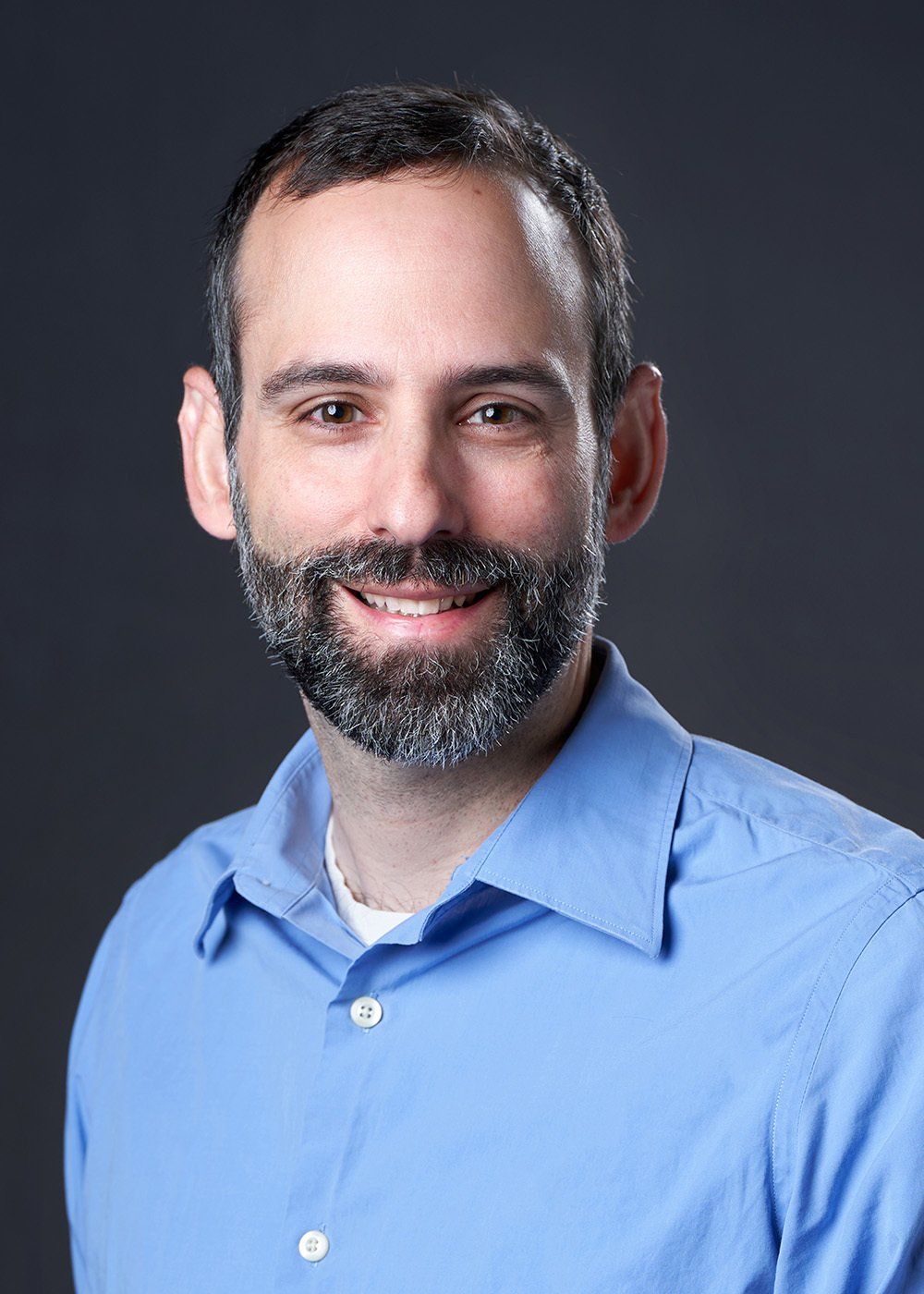 A man with a beard is wearing a blue shirt and smiling for the camera.