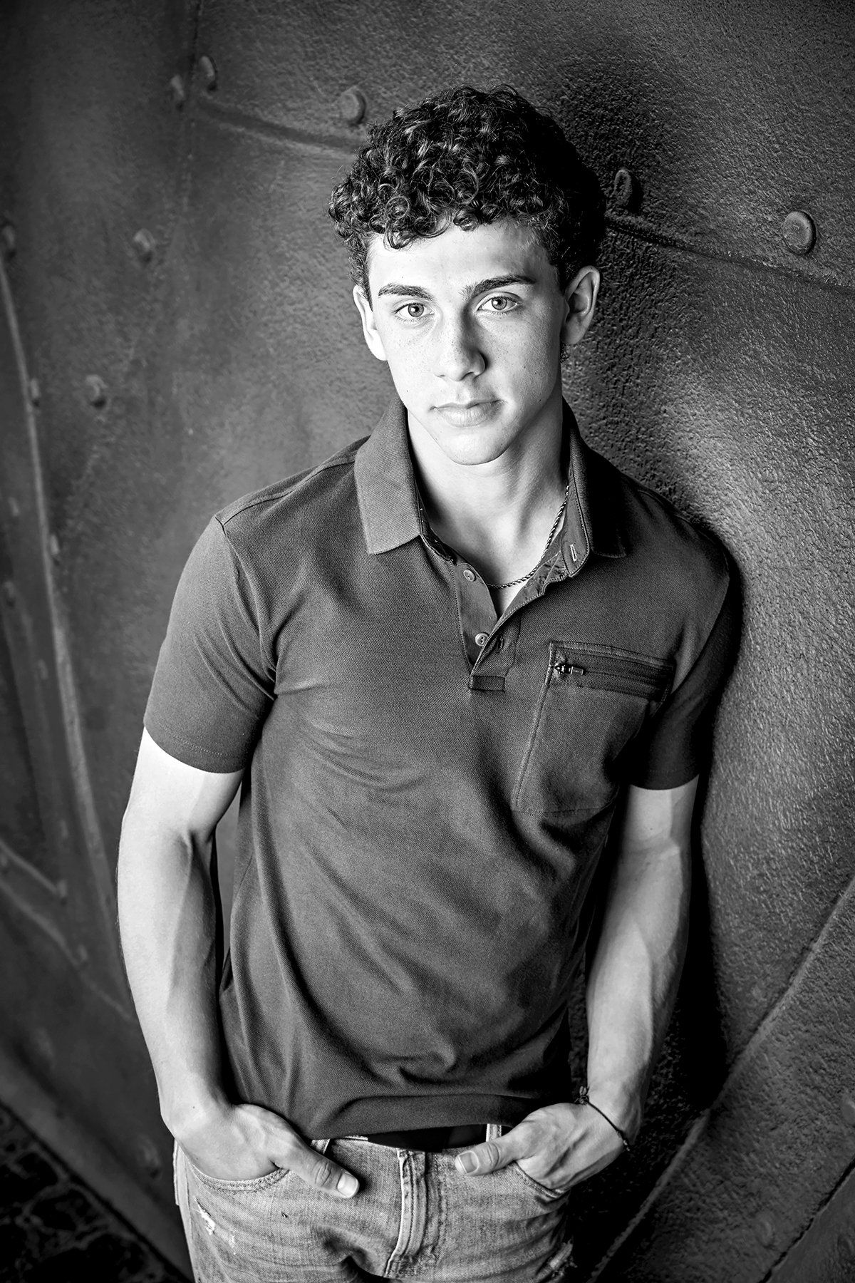 A young man is leaning against a wall with his hands in his pockets.