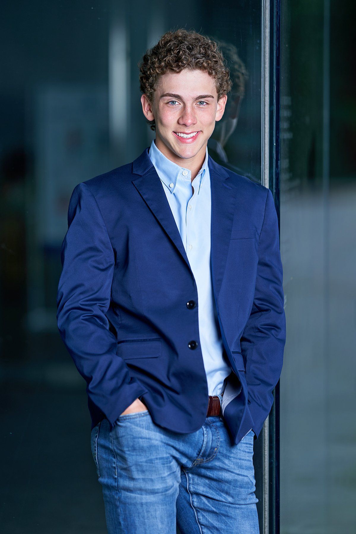 A young man in a blue jacket and jeans is standing in front of a glass door.