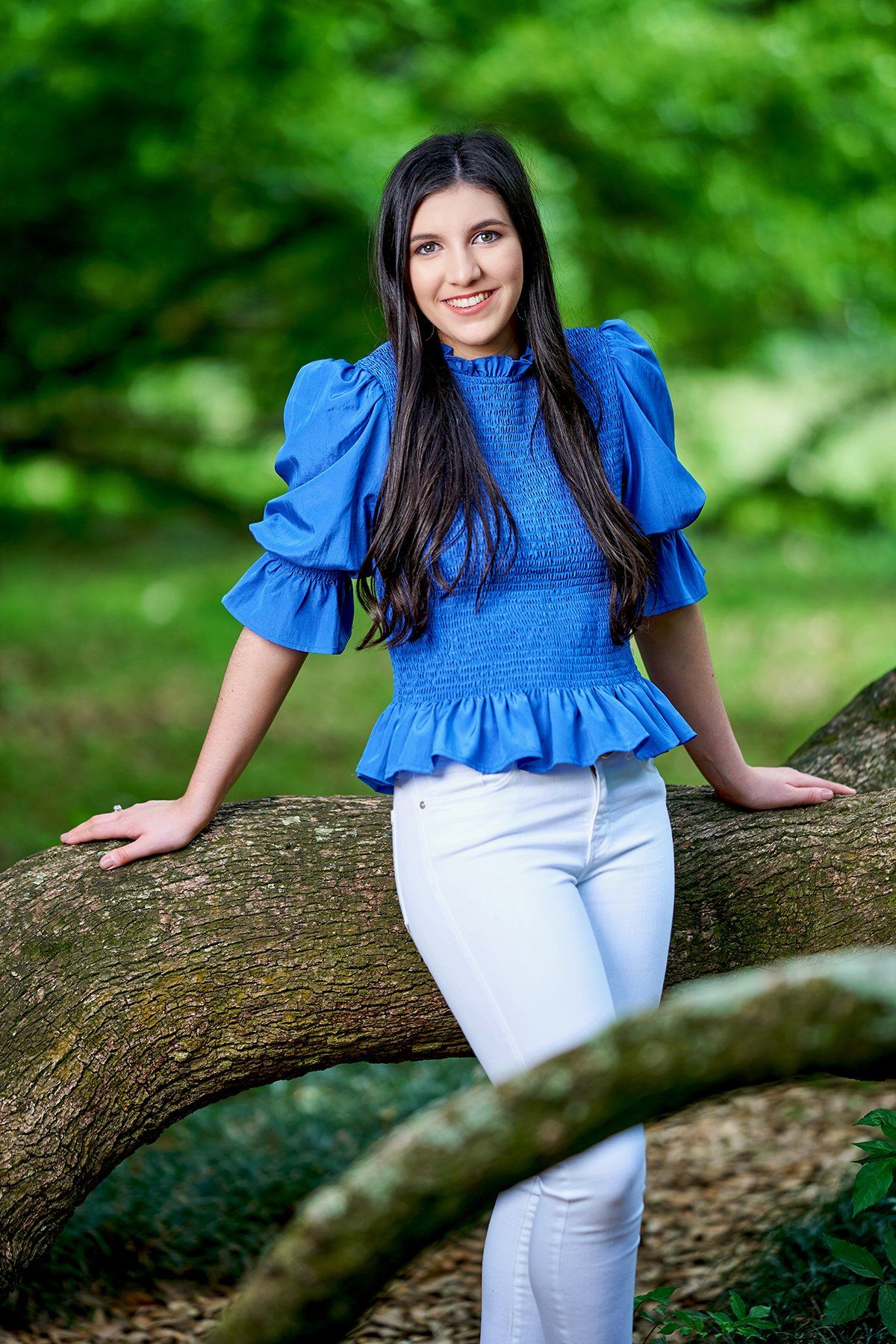 A woman in a blue top and white pants is leaning on a tree branch.