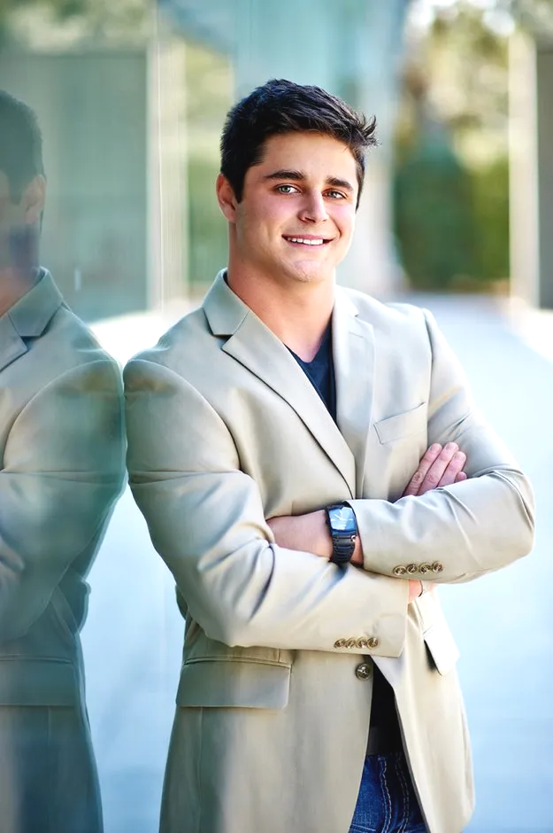 A young man in a tan suit is standing with his arms crossed.