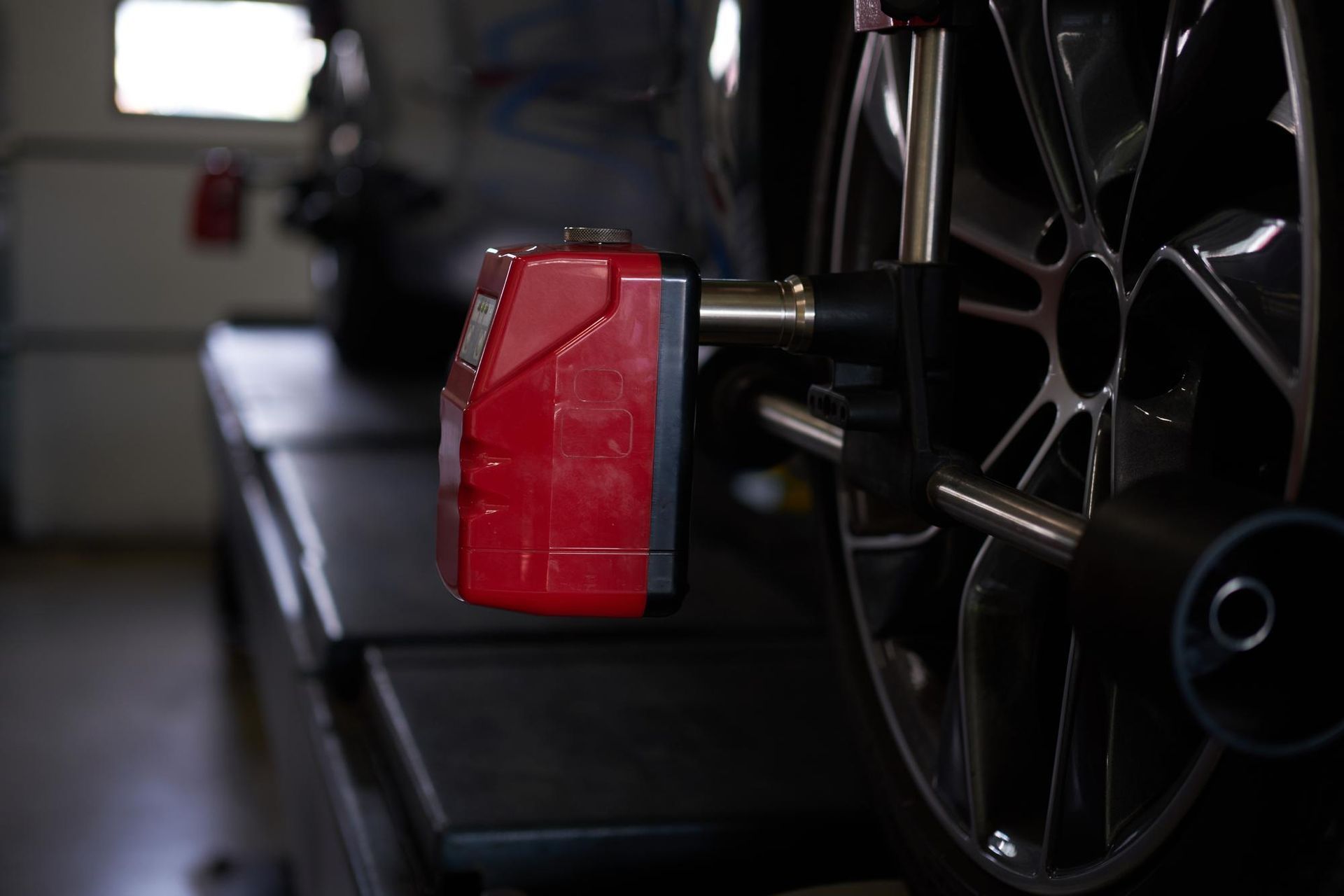 Wheel alignment machine attached to a car's wheel, likely in a garage. Red sensor, metallic wheel.