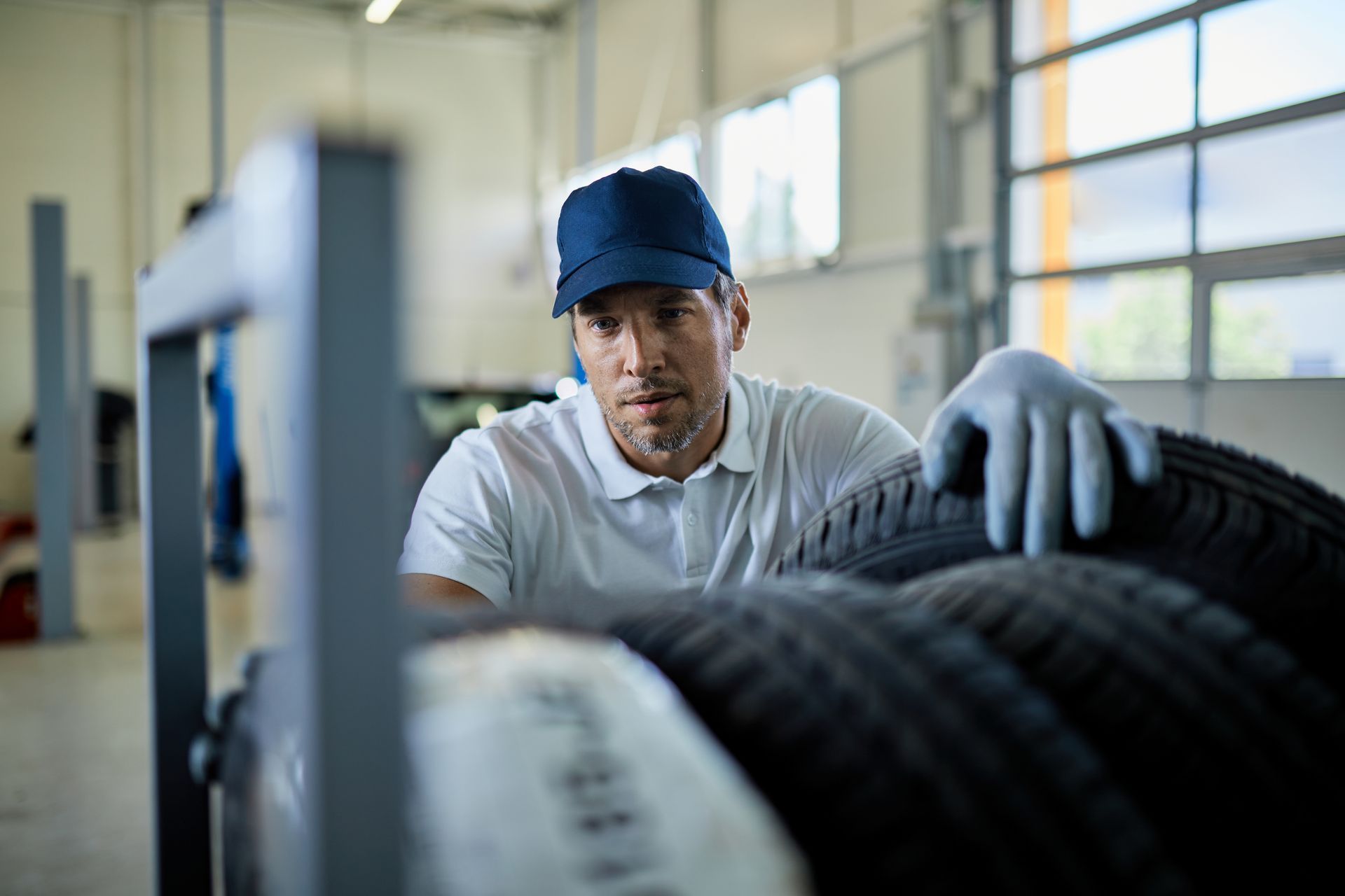 Mechanic in blue hat and gloves inspecting tires on a rack in a shop.