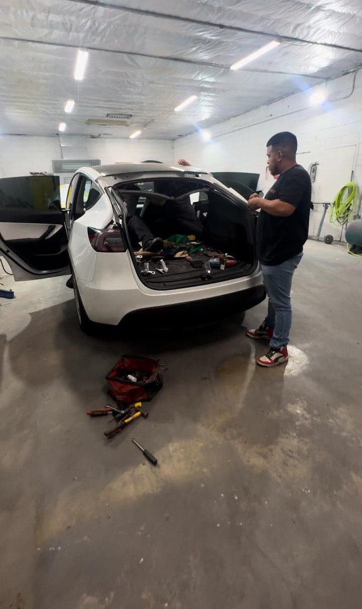Man working on the open trunk of a white car inside a workshop.