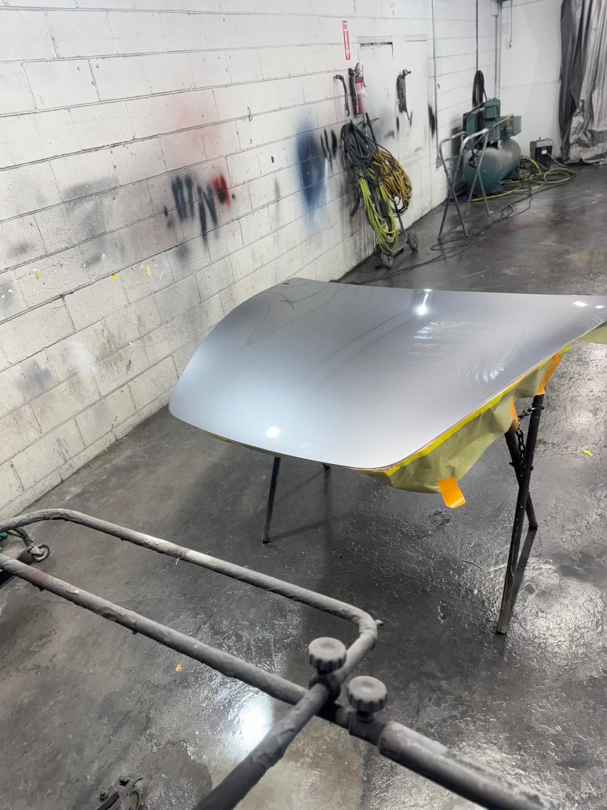 Car hood painted silver, on a stand in a workshop, near a spray painted wall.