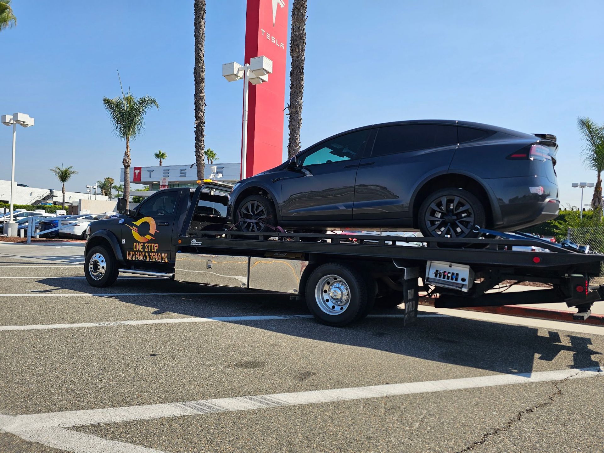 A dark gray Tesla Model X on a tow truck in front of a Tesla dealership under a blue sky.